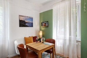A room with a wooden table, two chairs, a painting, a shelf with books, and a window with curtains. - Apartment 'Appartement In Historischem Haus' with Private Terrace, Private Garden and Wi-Fi (Ottersweier)