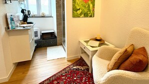 A compact living space with a kitchenette, a dining area, a sofa with cushions, a patterned rug, and a painting of a flower. - 'Single Room Ahorn, Nature, Wellness' with Private Terrace, Garden, and Wi-Fi (Ottersweier)