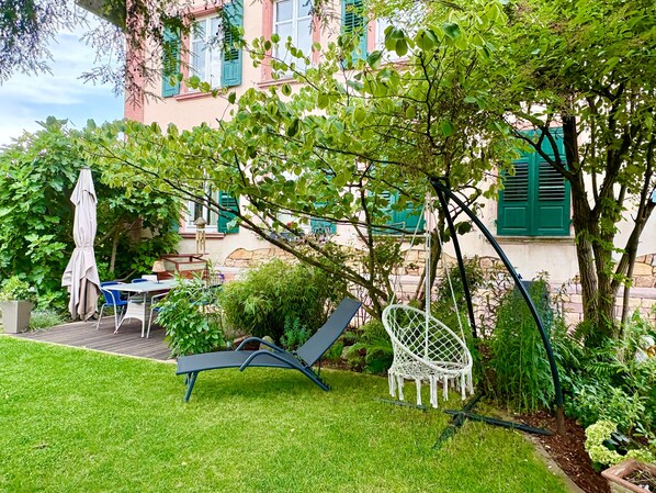 A garden with a lawn, a table, chairs, a hanging chair, and a building with green shutters in the background.