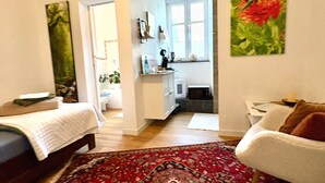 A room with a bed, a red patterned rug, a white chair, a kitchen area, and a painting of a flower. - 'Single Room Ahorn, Nature, Wellness' with Private Terrace, Garden, and Wi-Fi (Ottersweier)