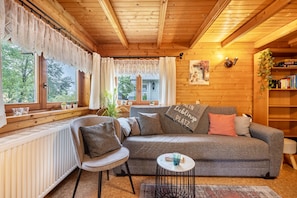 Ein gemütliches Zimmer mit Holzbalkendecke, einem grauen Sofa mit Kissen, einem kleinen runden Tisch und einem Bücherregal mit Büchern und einer Pflanze.