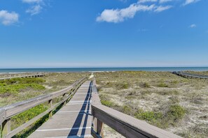 Ein hölzerner Steg führt zu einem Strand mit Dünen und blauem Himmel.