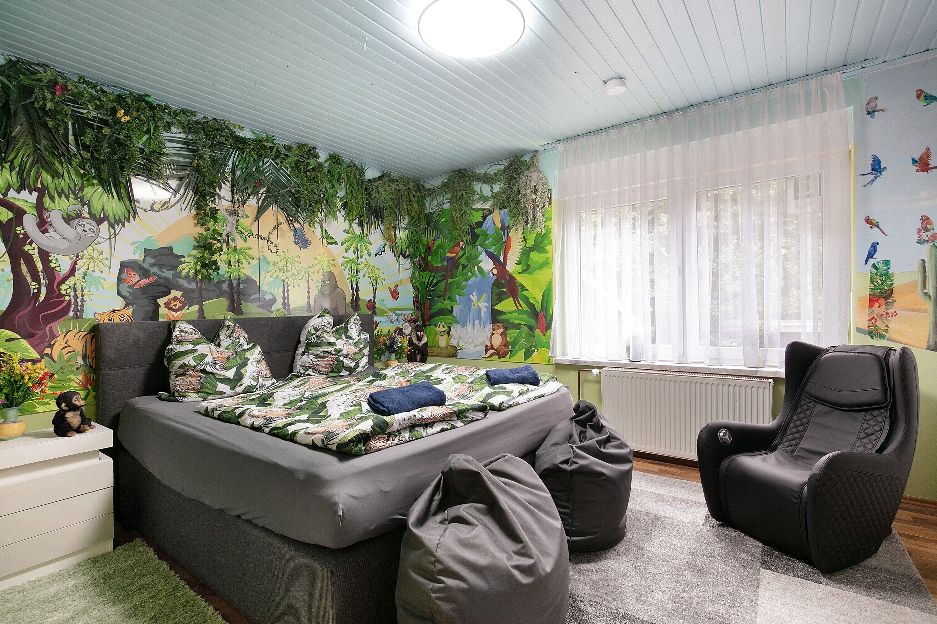Une chambre à coucher comprenant un lit, un fauteuil de massage, des poufs et une fresque murale représentant une scène de jungle.