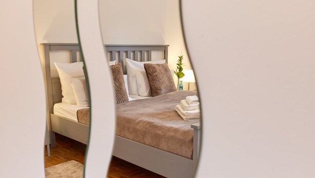 Een opgemaakt bed met kussens, een nachtkastje met een lamp, en een houten vloer.