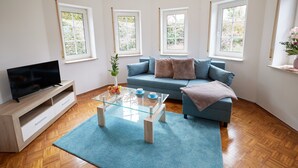 Een moderne woonkamer met een bank, een glazen salontafel, een tv op een houten dressoir en een raam met uitzicht op groen.
