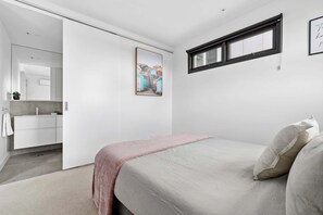 Un dormitorio con cama, ventana, un cuadro y baño con lavamanos y espejo.