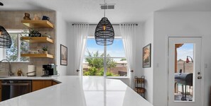 Une cuisine moderne avec des plans de travail blancs, des étagères en bois et une grande fenêtre agrémentée de rideaux.