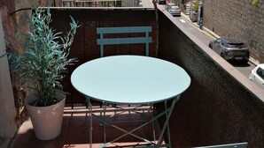 Ein Balkon mit rundem Tisch und zwei Stühlen, einem Blumentopf sowie Blick auf eine Straße mit geparkten Autos und Gebäuden im Hintergrund.