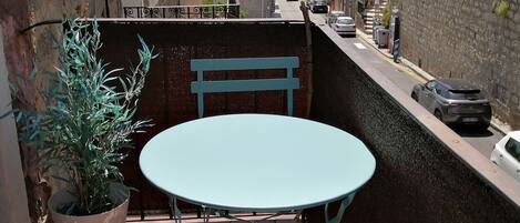 Un balcon avec une table ronde et deux chaises, un pot de fleurs et une vue sur une rue bordée de voitures garées, avec des bâtiments en arrière-plan.