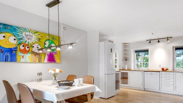Uma cozinha moderna com armários brancos, um espaço de refeições com mesa branca e cadeiras marrons, e um mural colorido na parede.