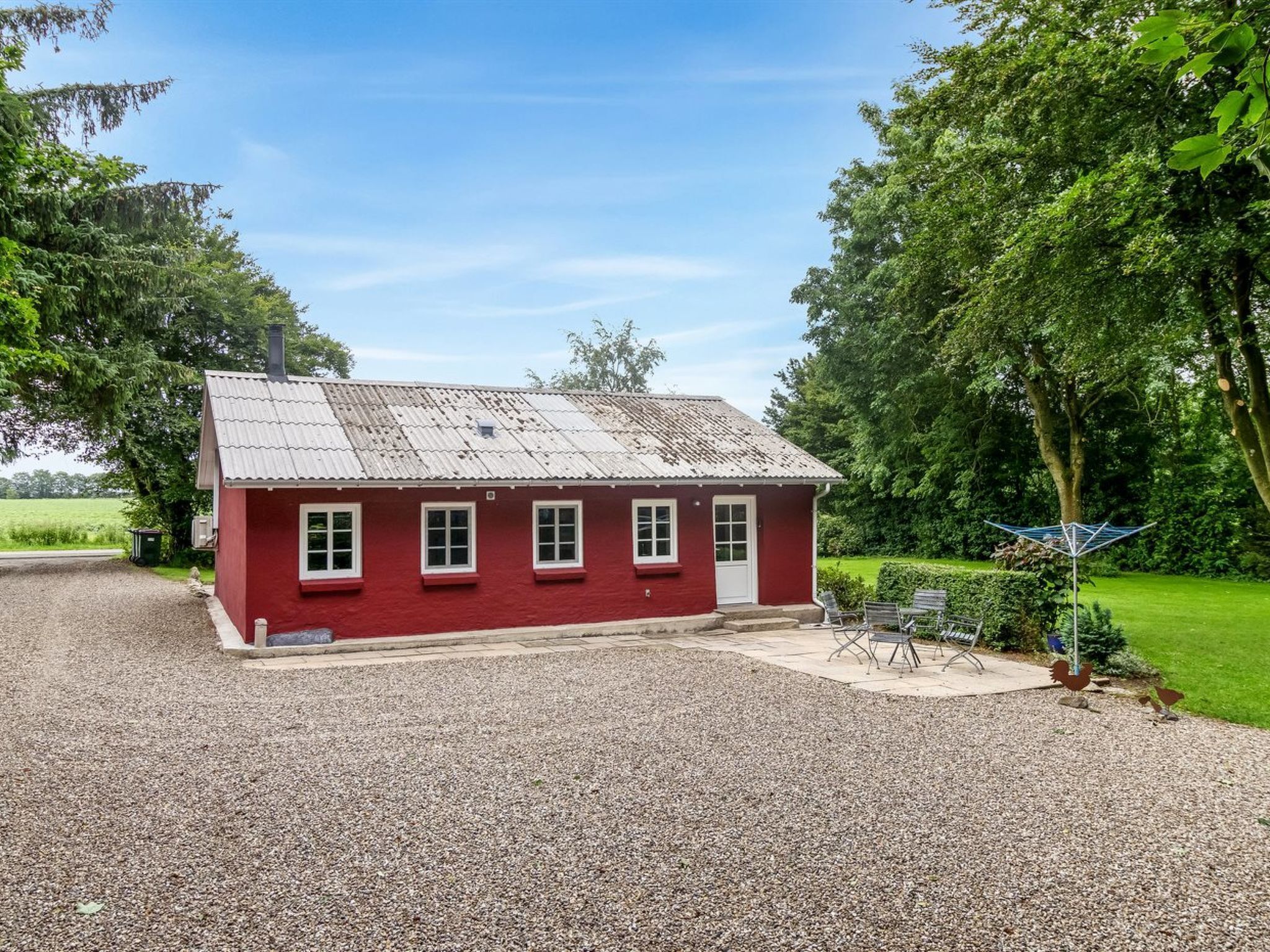 Una casa a un piano di colore rosso con vialetto sterrato e un piccolo patio con sedie e tavolo in metallo.