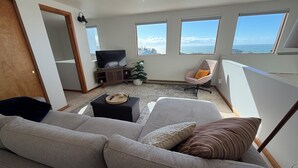 A modern living room with a grey sectional sofa, a small black ottoman, a wooden entertainment unit, a potted plant, and a chair with a cushion. Large windows offer a view of the sea.