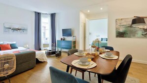 A modern hotel room with a bed, a dining area with a table set for two, a TV, and artwork on the walls. - Splendid Apartment in the Heart of the Chic 16th (Paris)