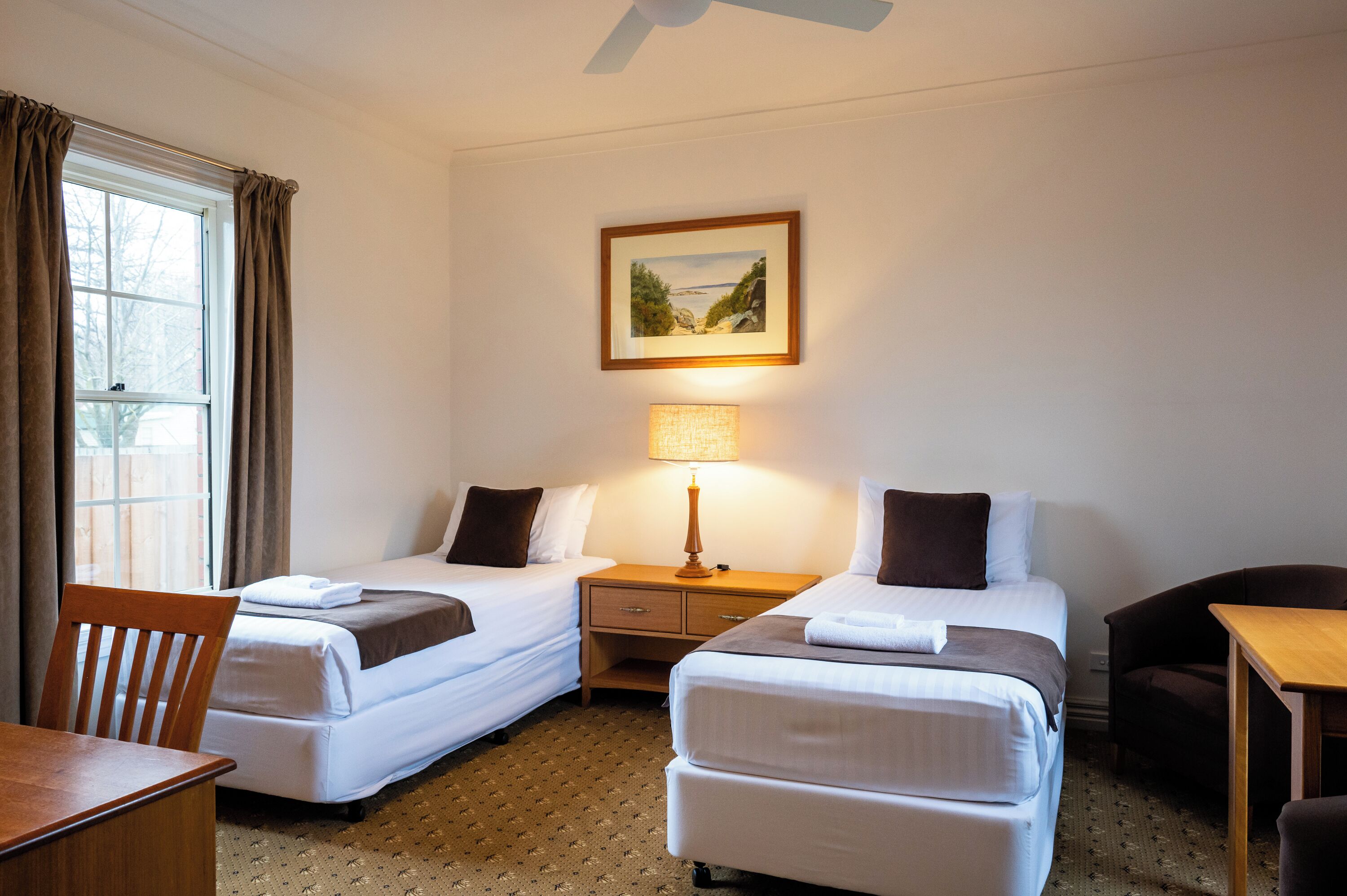 A hotel room with two single beds, a desk, a chair, a lamp, and a framed picture on the wall.