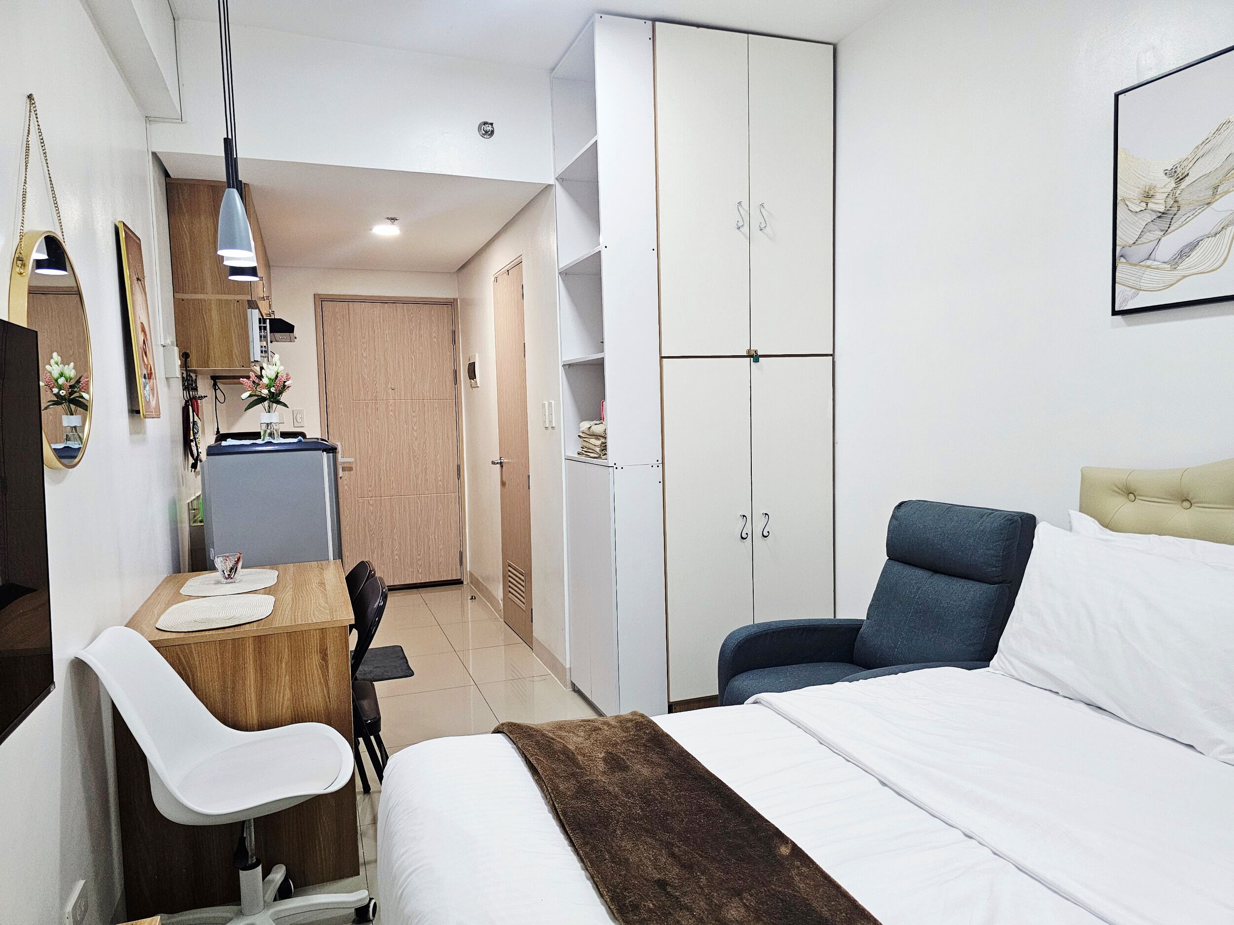 A bedroom with a bed, a desk, a chair, a wardrobe, and a kitchenette.