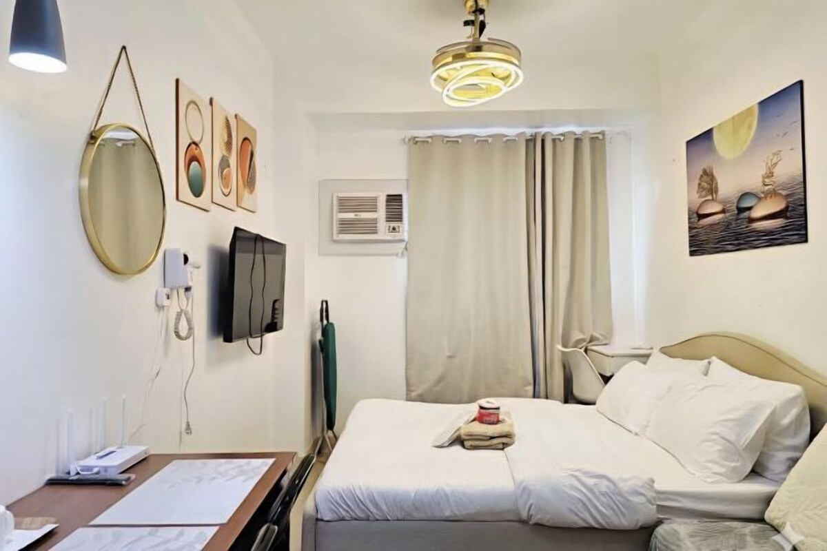 Habitación de hotel con cama, escritorio con silla, televisión de pantalla plana, espejo redondo, obras de arte en las paredes y lámpara en el techo.