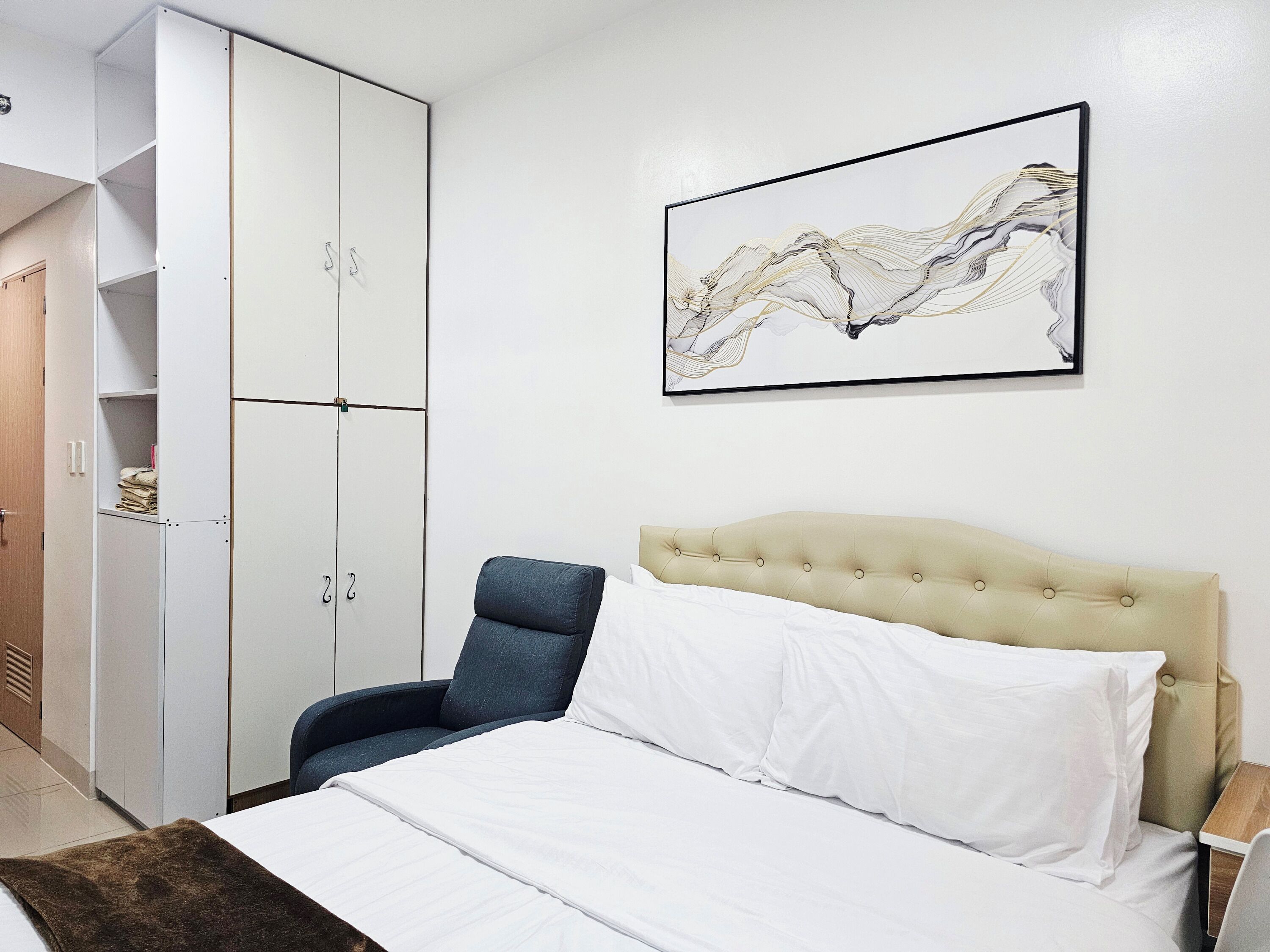 A bedroom with a bed, a chair, a wardrobe, and a wall-mounted artwork.