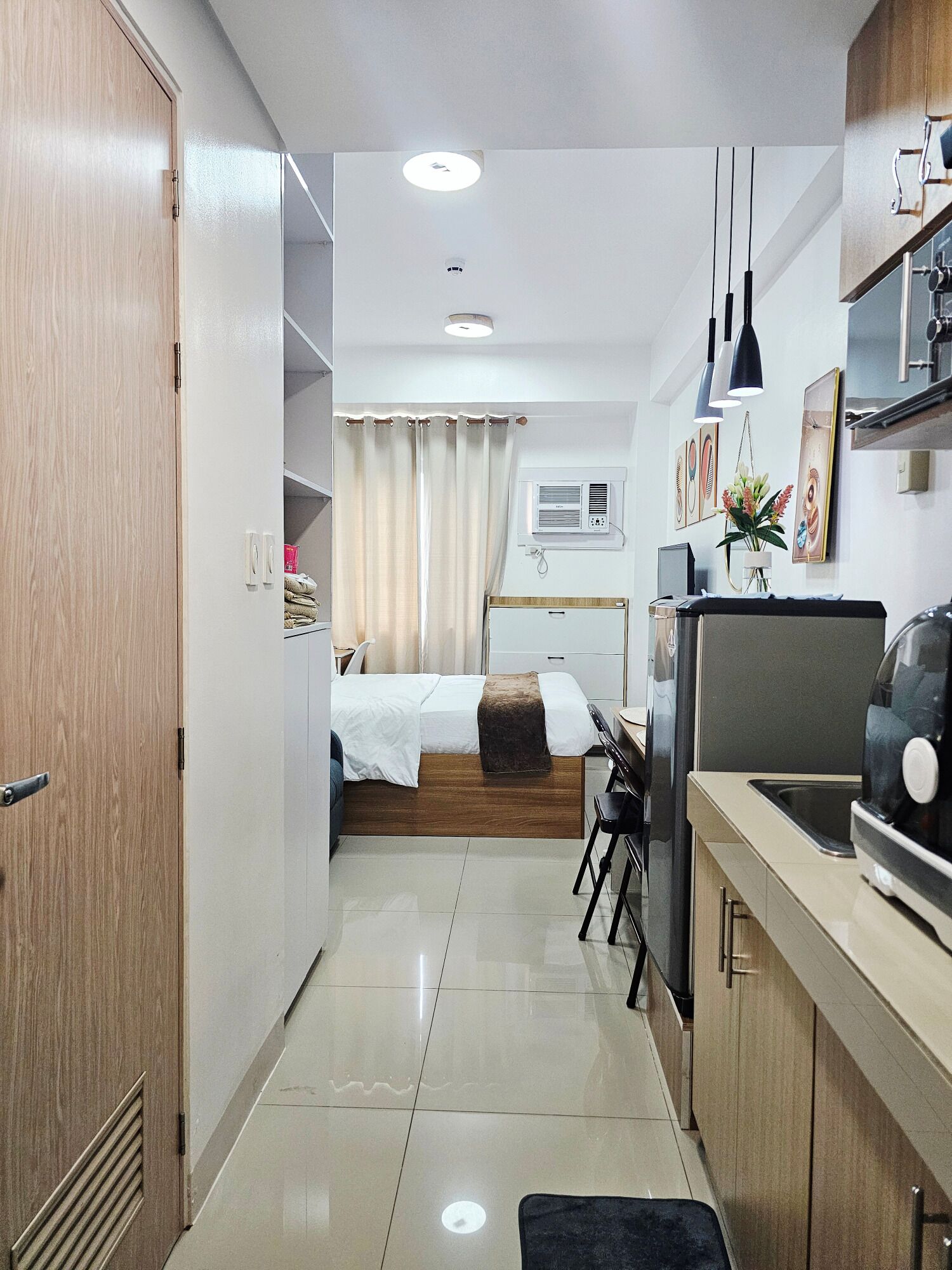 A compact hotel room with a kitchenette, a bed with a wooden headboard, a small desk, and a bathroom with a shower.