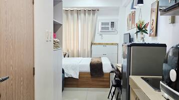 A compact hotel room with a kitchenette, a bed with a wooden headboard, a small desk, and a bathroom with a shower.