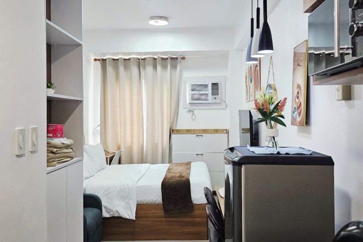 Habitación de hotel pequeña con cama, escritorio, refrigerador y ventana con cortinas.