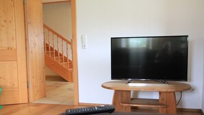 Un soggiorno con televisione su un mobile in legno, telecomando sul divano e una scala in legno sullo sfondo.