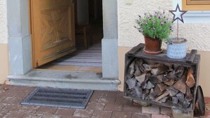 Una porta in legno con pannelli di vetro, una pianta in vaso con fiori viola e una pila di legna davanti a un edificio con un muro strutturato e una finestra.