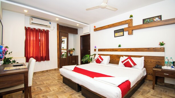 A hotel room with a large bed, wooden headboard, a desk with a chair, a small table with a phone and water bottles, and a ceiling fan. - Octave Verdisso Hotel (Bengaluru)