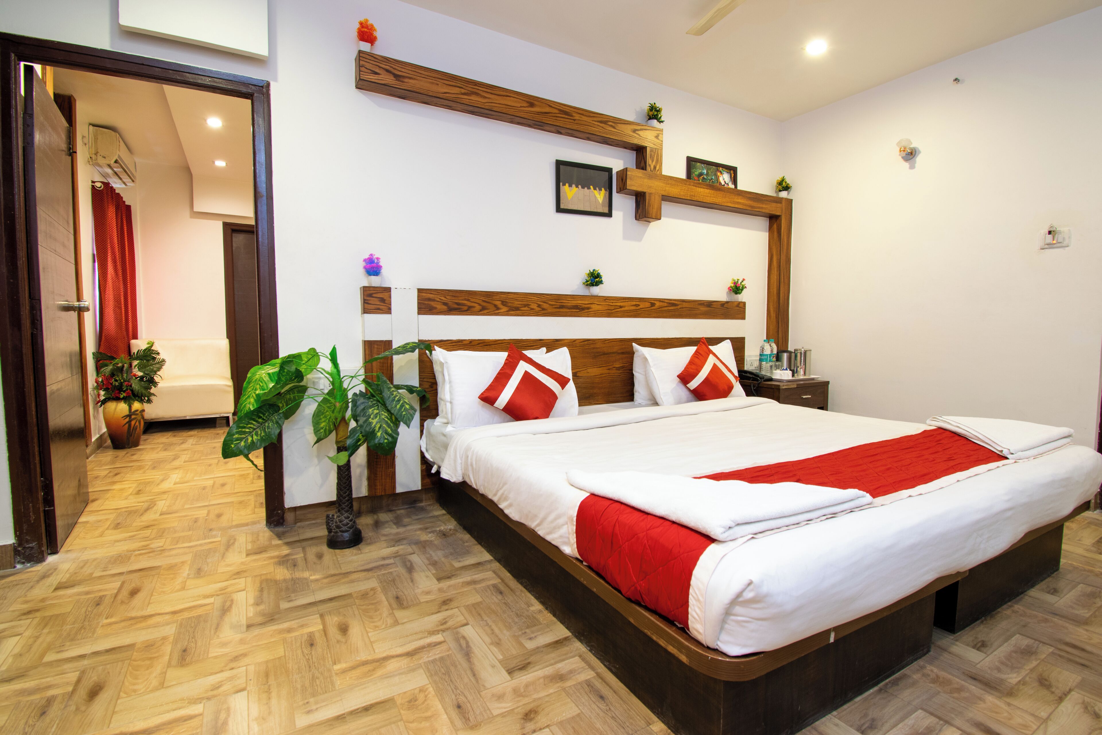 A hotel room with a wooden headboard, a large bed with white bedding and red accents, a bedside table with a lamp, and a potted plant.