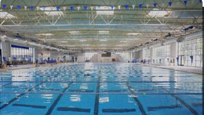 Une piscine intérieure dotée de plusieurs couloirs, d’un plafond haut avec des poutres apparentes et de grandes fenêtres sur un des côtés.