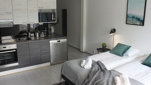 Ein modernes Hotelzimmer mit Küchenbereich, einem Bett mit Handtüchern, einem Nachttisch mit Lampe sowie einem Bild an der Wand.