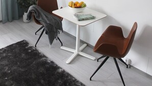 Ein moderner Essbereich mit einem weißen Tisch, zwei braunen Stühlen, einer Obstschale und einer grauen Decke auf einem Stuhl.