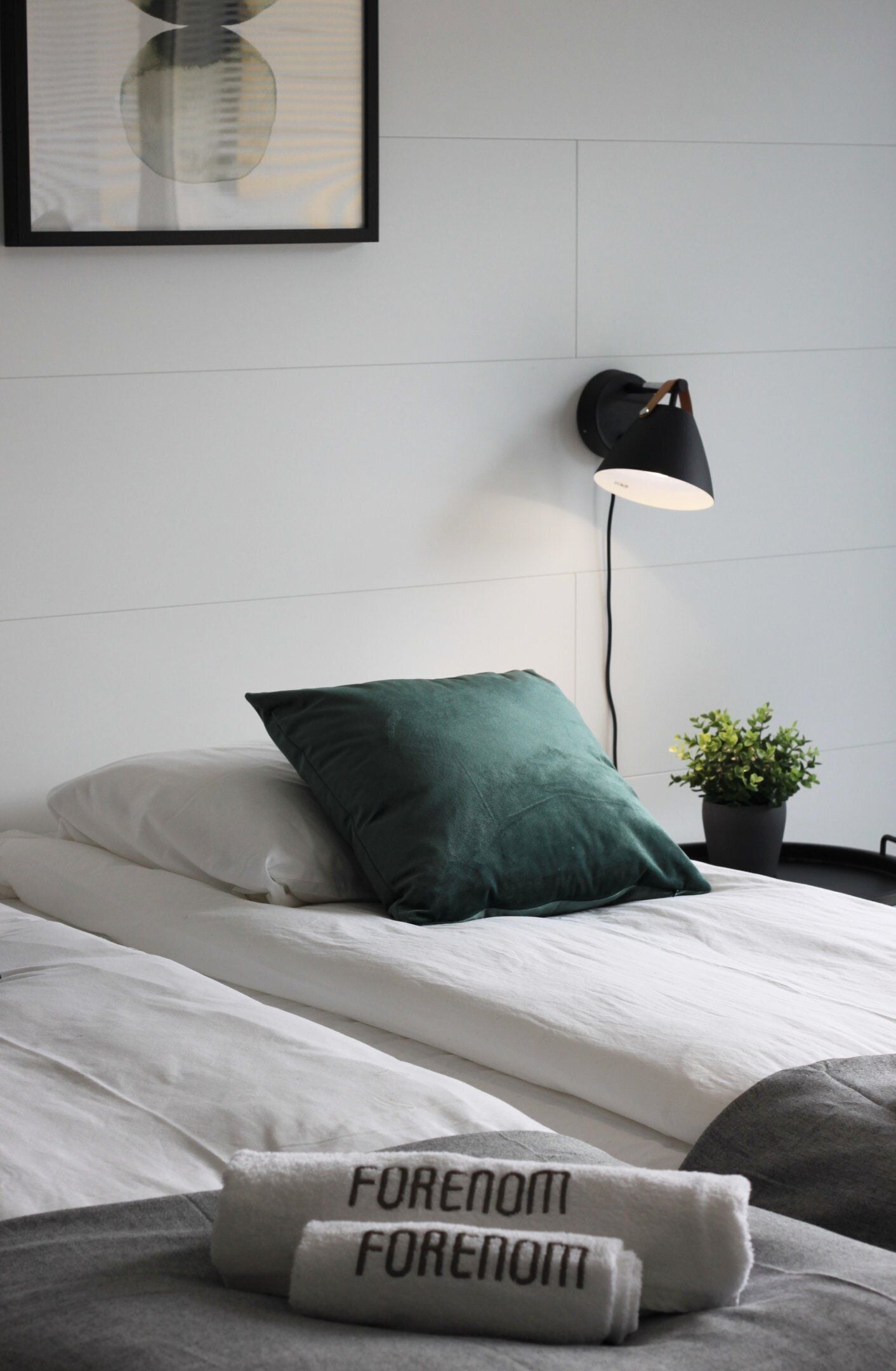 Una cama bien hecha con ropa blanca y una almohada verde, una pequeña planta en maceta y un cuadro en la pared.