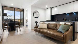 Uma sala de estar moderna com sofá de couro, uma área de jantar com mesa redonda e uma cozinha com armários brancos e bancada preta.