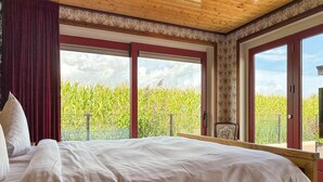 A bedroom with a large bed, two windows with red frames, and a view of a cornfield outside.