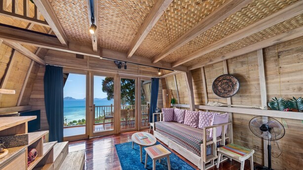 A cozy, wooden interior with a large window offering a view of the sea, a sofa, a small table, and a fan.
