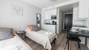 A modern bedroom with two beds, a small bedside table, a nightstand, a desk with a chair, and a kitchenette. - Forenom Serviced Apts Vantaa Koivuhaka (Vantaa)