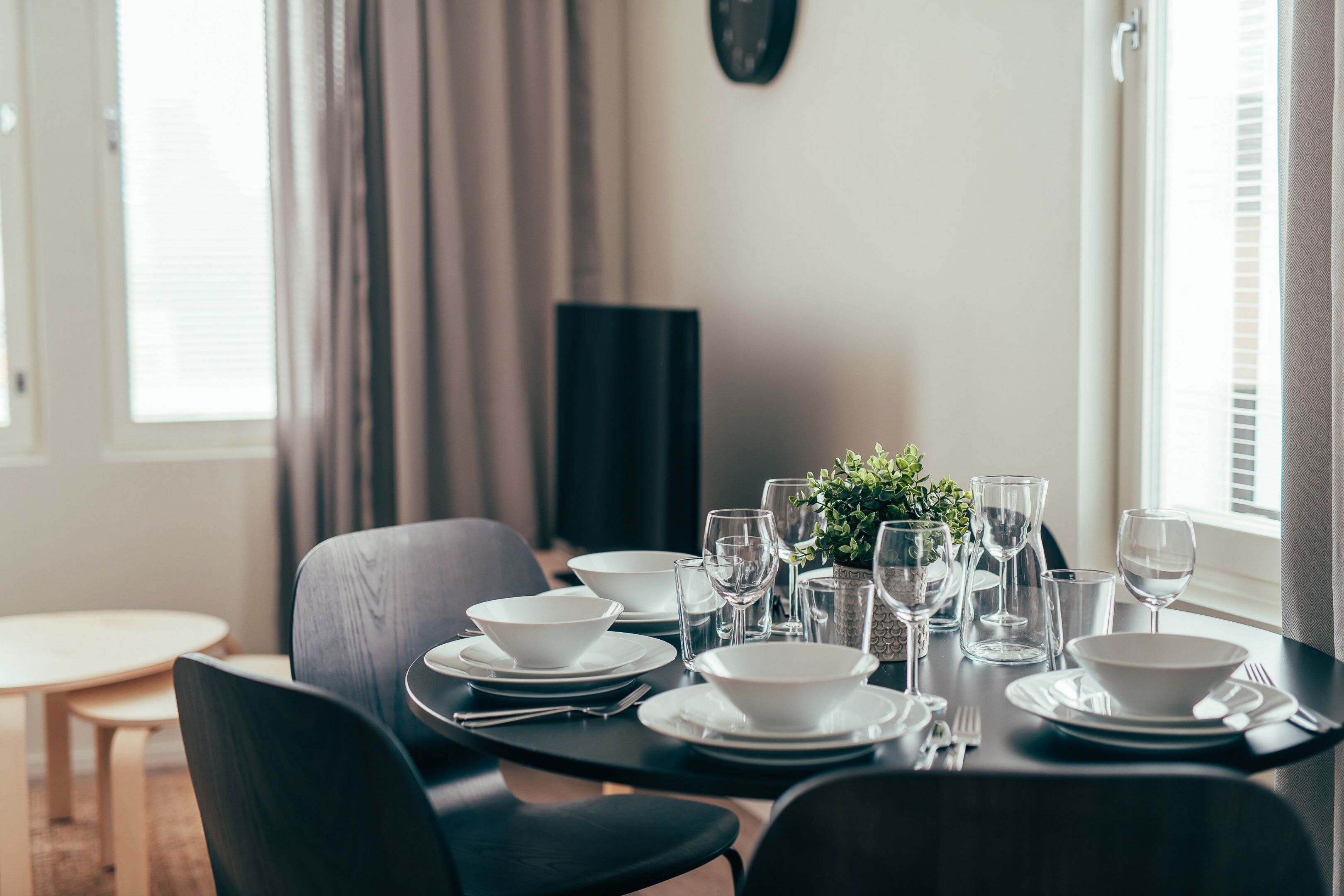 Una mesa de comedor dispuesta con cuencos, platos y vasos blancos, rodeada de sillas oscuras.