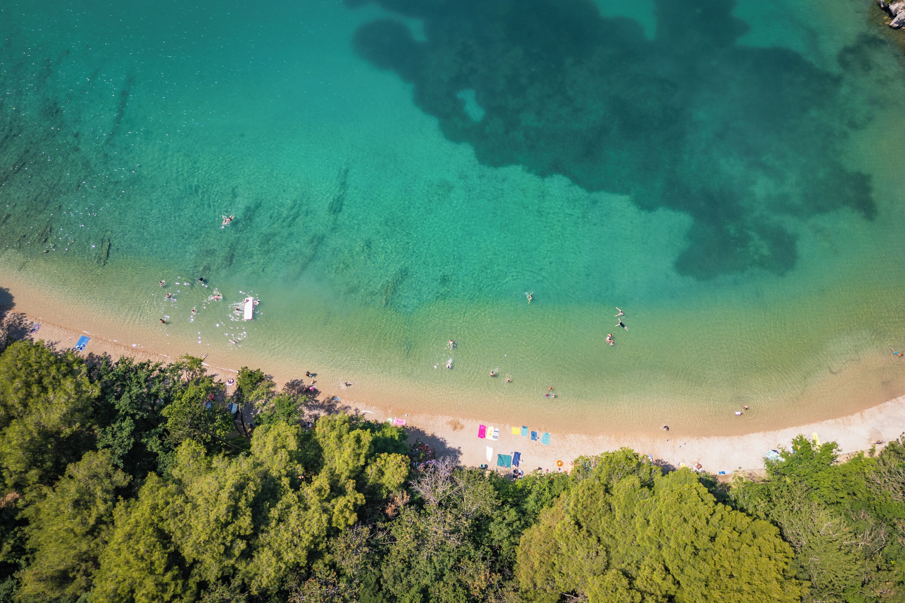 An aerial view of a beach with clear turquoise waters, a sandy shore, and dense green trees.