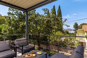 Un balcon avec des sièges d’extérieur et une table dressée, donnant sur un paysage arboré et offrant une vue lointaine sur une étendue d’eau.