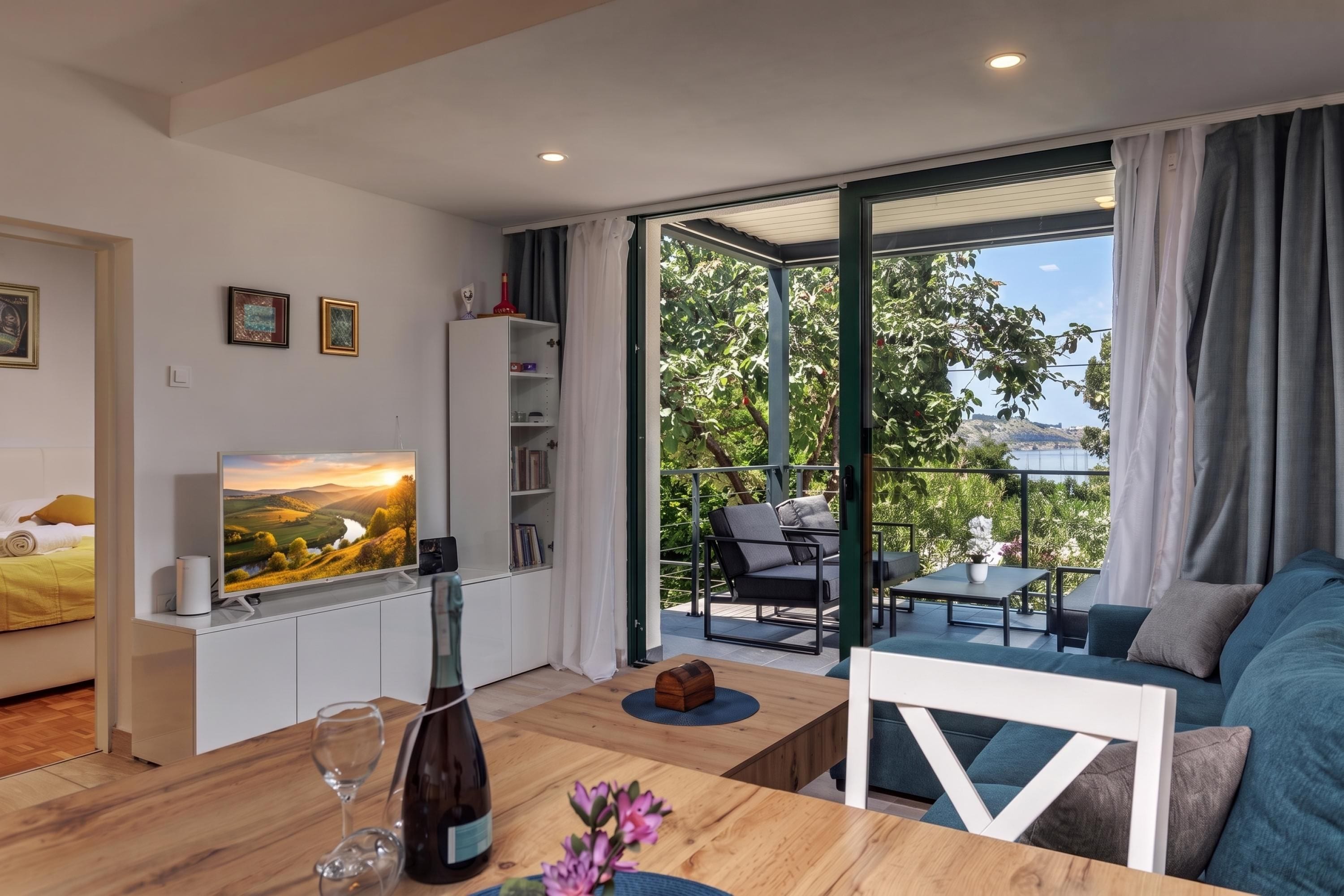 Un salon moderne avec une télévision, une table basse en bois et un canapé. Il y a un balcon avec du mobilier d’extérieur et une vue sur la verdure.