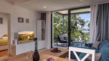A modern living room with a television, a wooden coffee table, and a sofa. There is a balcony with outdoor furniture and a view of greenery.