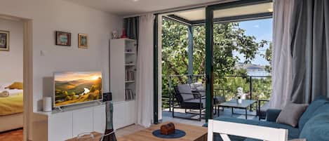 A modern living room with a television, a wooden coffee table, and a sofa. There is a balcony with outdoor furniture and a view of greenery.