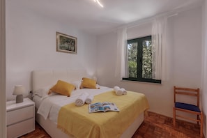 A neatly arranged bedroom with a bed, a bedside table, a chair, a window with curtains, and a framed picture on the wall. - Beachside House Lara (Omisalj)