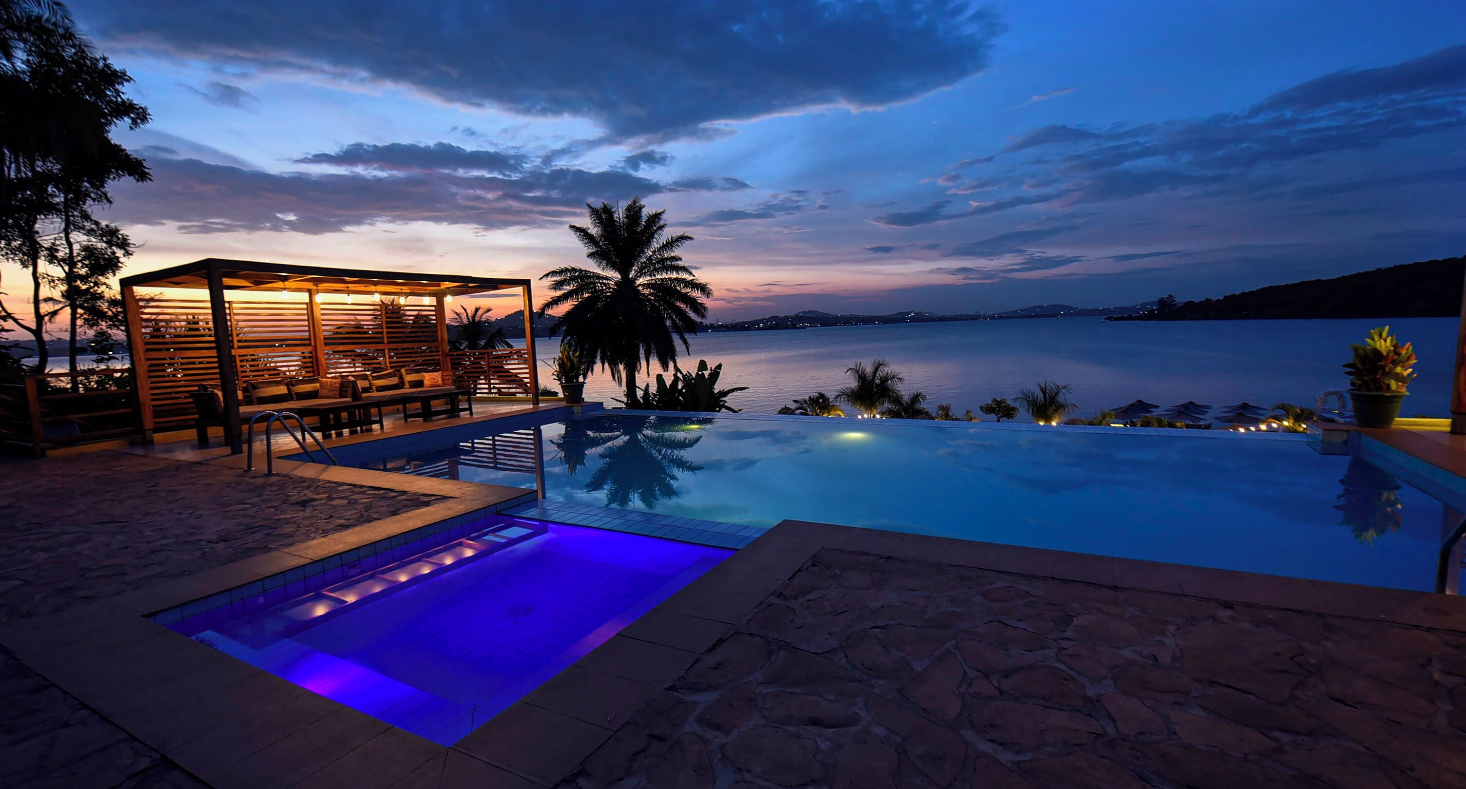A swimming pool with a wooden deck and lounge area, surrounded by palm trees and a view of the water.