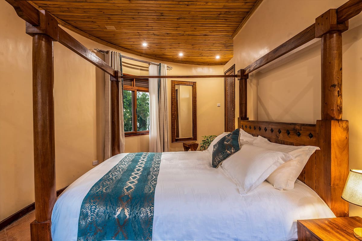 A bedroom with a wooden ceiling, a large bed with white and blue bedding, a nightstand, and a window with curtains.