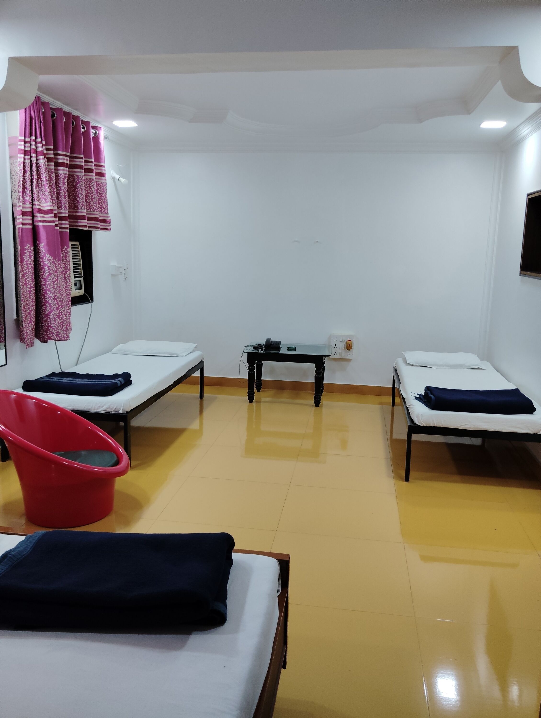 A room with two single beds, a red chair, a small table, and a wall-mounted heater.