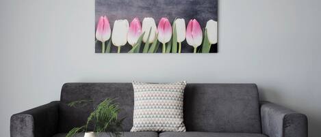 A grey sofa with a patterned pillow, a black coffee table with a book titled "ATHENS RIVIERA," and a wall-mounted painting of pink and white tulips.