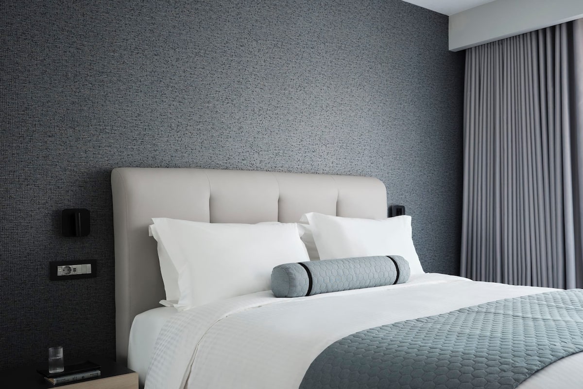 Una habitación de hotel moderna con una pared gris con textura, una cama grande con ropa de cama blanca y gris, una mesita de noche de madera con un vaso y un objeto negro encima.