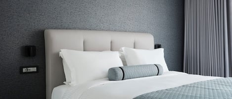 A modern hotel room with a grey textured wall, a large bed with white and grey bedding, a wooden nightstand with a glass, and a black object on it.