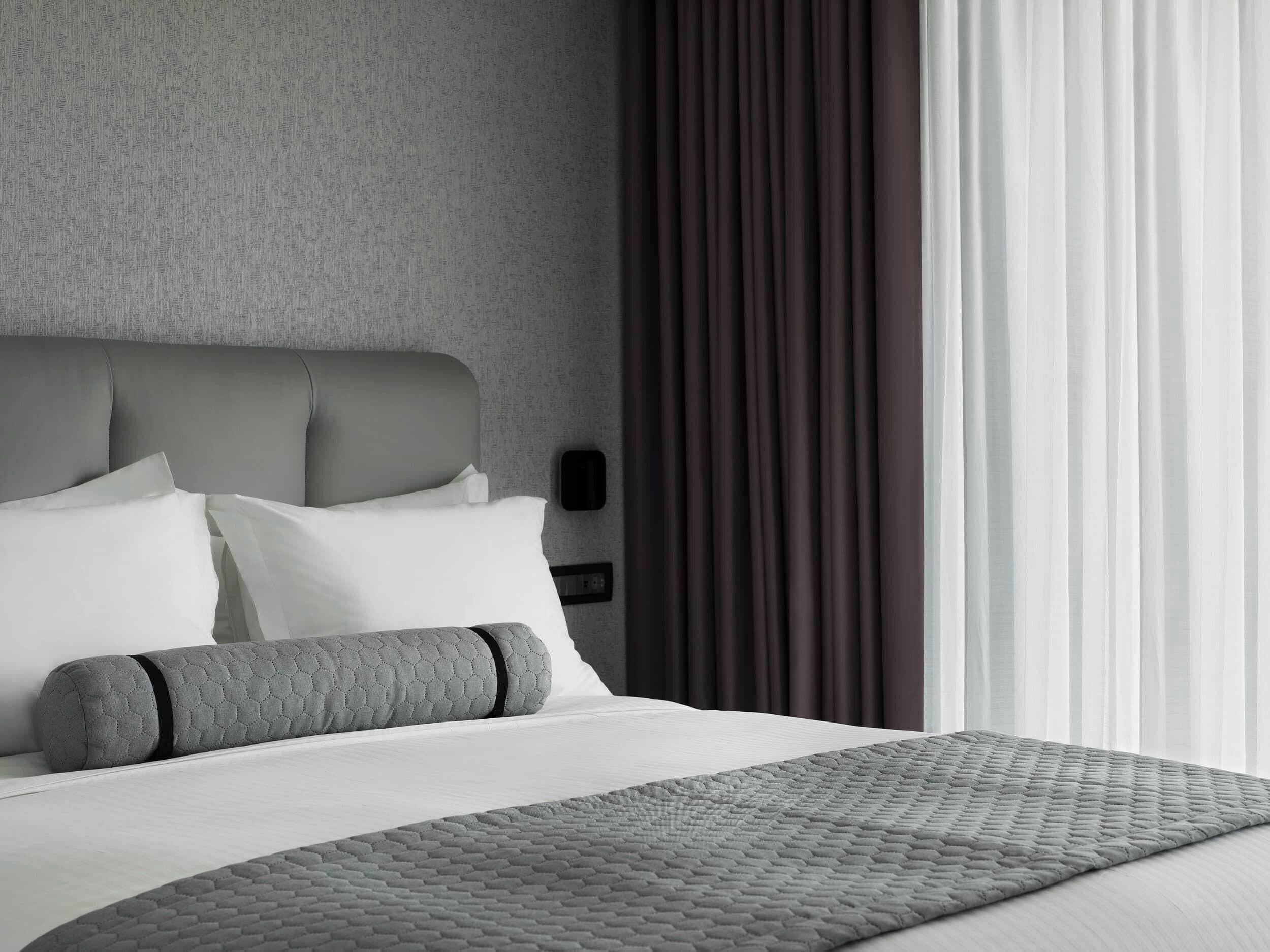 A modern hotel room with a grey headboard, white bedding, and a grey quilted bedspread.